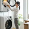 明るいランドリールームで、縦型洗濯機の上に設置された乾太くんに洗濯物を入れている日本人女性。横には泥汚れのついた子供服の山。