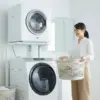 洗濯機の上部にガス衣類乾燥機が設置された洗面所で、洗濯カゴを持って笑顔で次の洗濯準備をする日本人女性の様子。洗濯と乾燥の並列処理による時短イメージ。