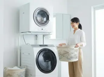 洗濯機の上部にガス衣類乾燥機が設置された洗面所で、洗濯カゴを持って笑顔で次の洗濯準備をする日本人女性の様子。洗濯と乾燥の並列処理による時短イメージ。
